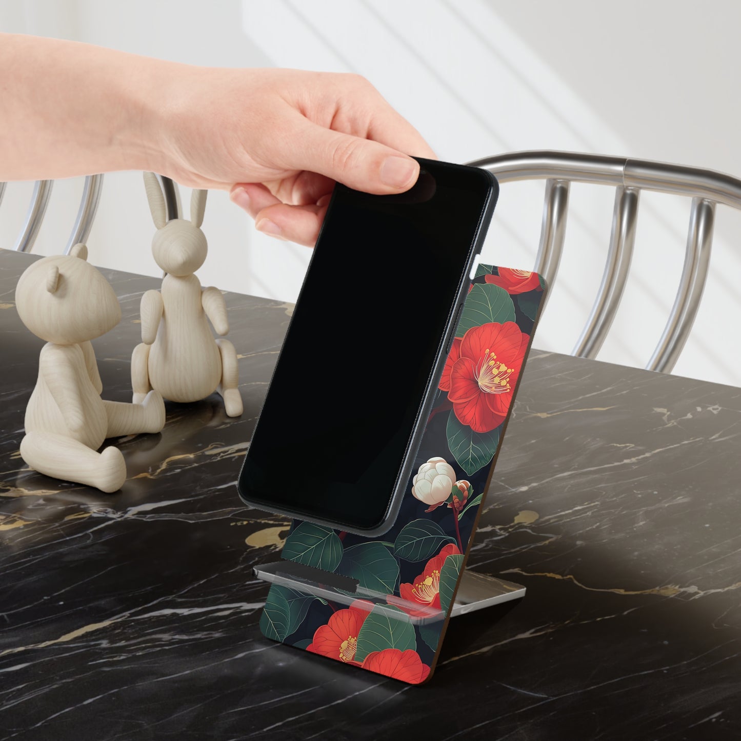 Phone on a floral stand being held by a hand on a marble surface with decorative items.