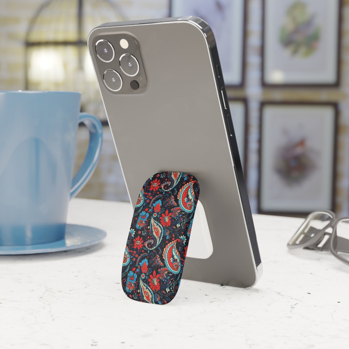 Phone stand with floral design holding a smartphone next to a blue mug on a table.