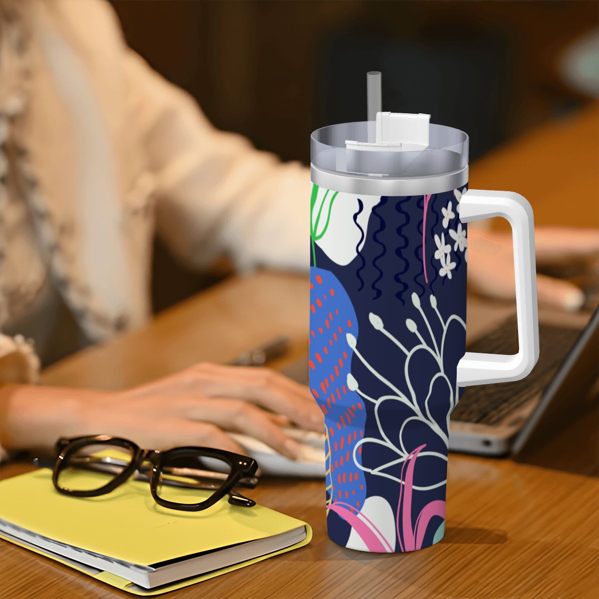 Colorful insulated mug with handle on a desk next to a person using a laptop.