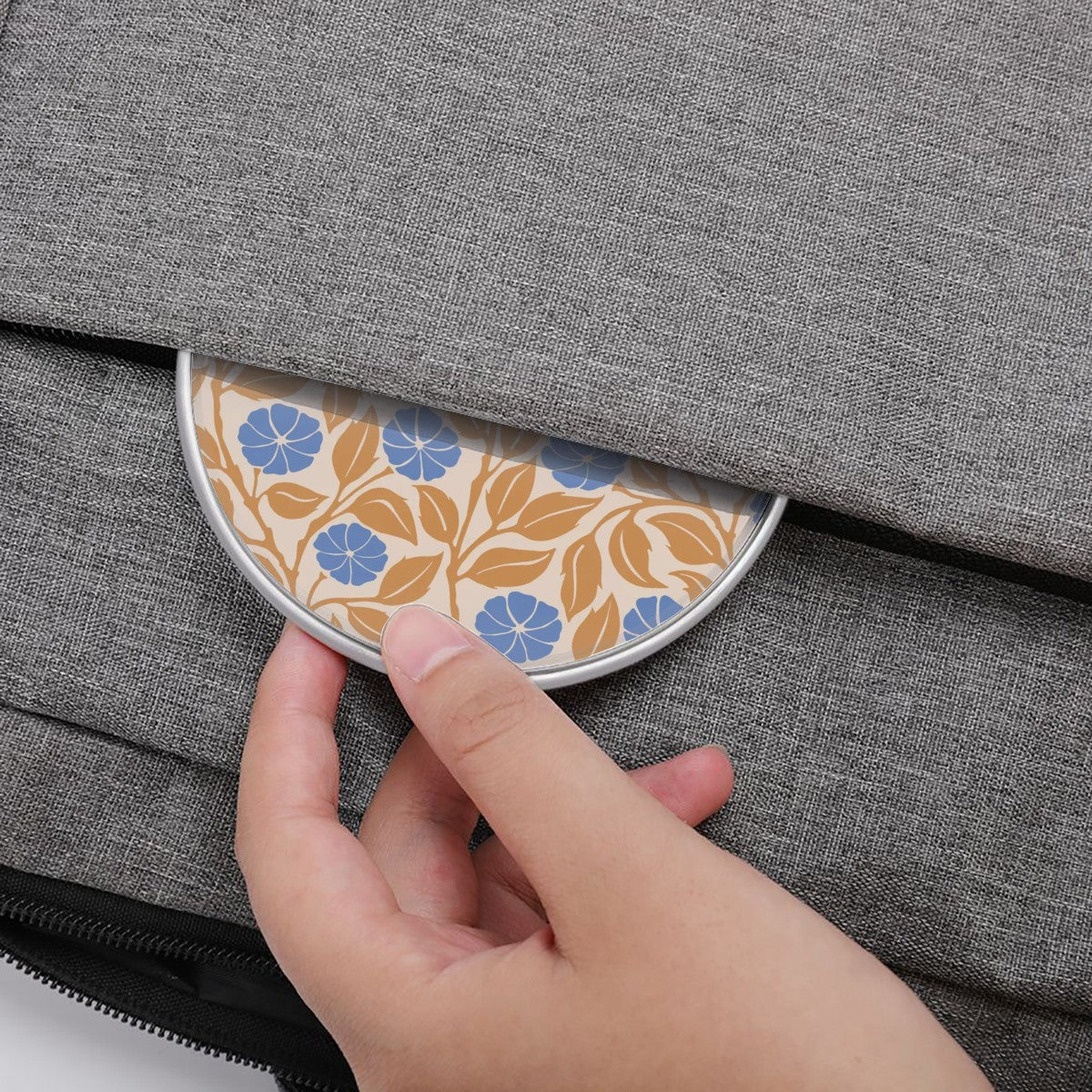A close-up of a hand pulling open a gray fabric pouch to reveal a round decorative tin with a blue and tan floral pattern.