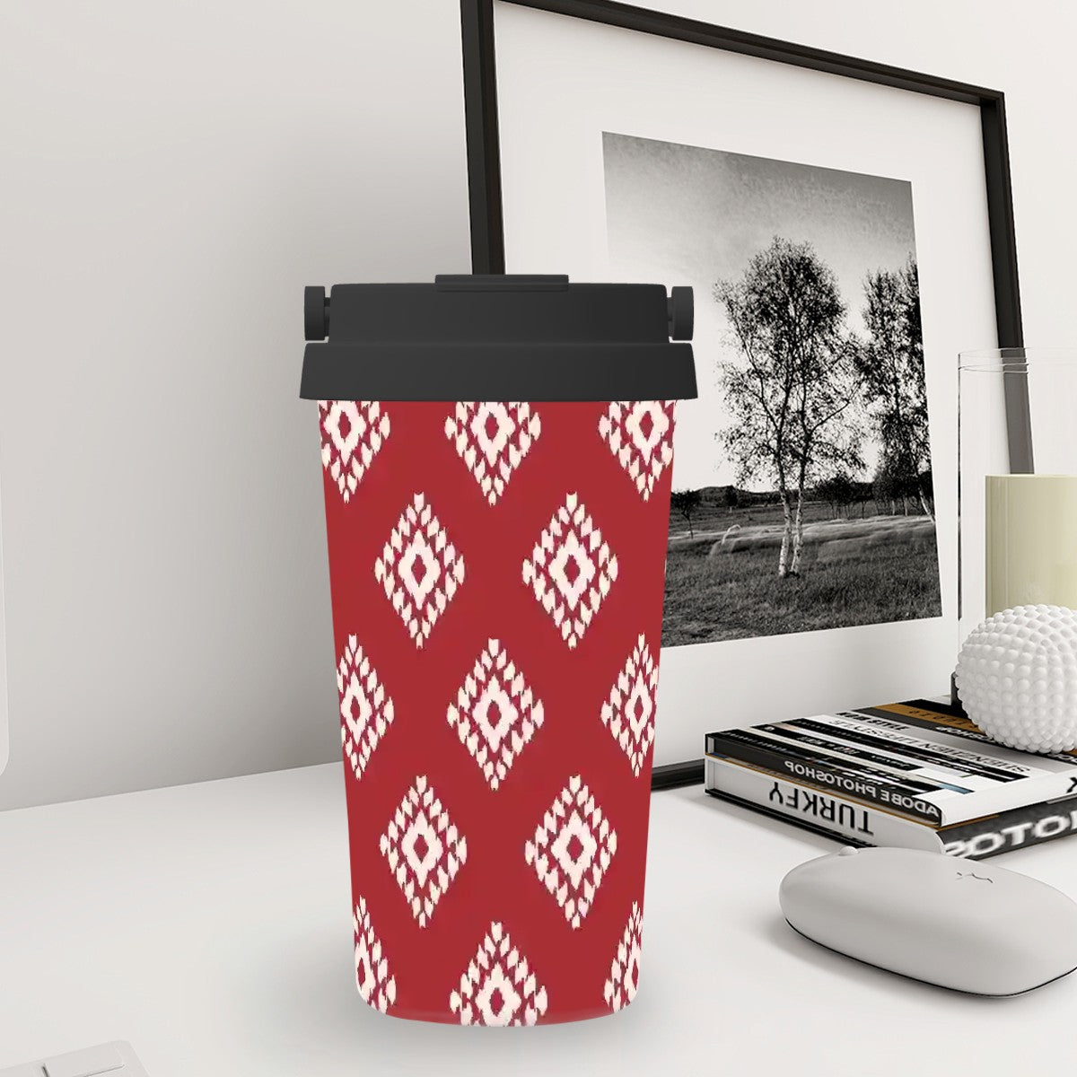 Red patterned tumbler with black lid on a desk next to a framed picture and books.