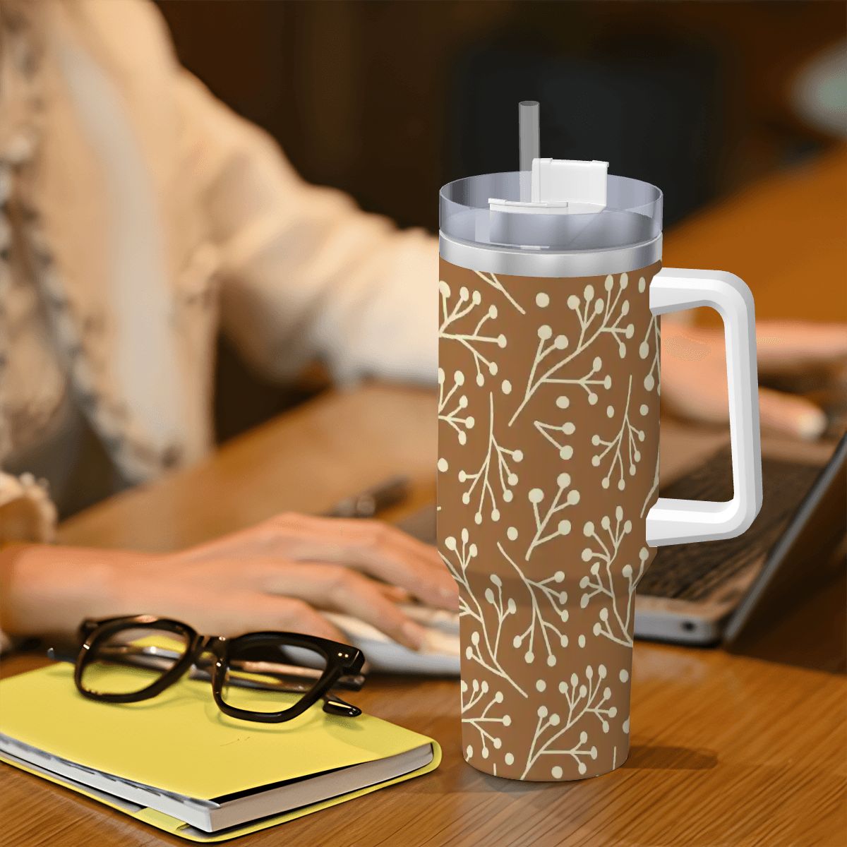 Brown tumbler with white floral pattern on a desk with a person using a laptop.