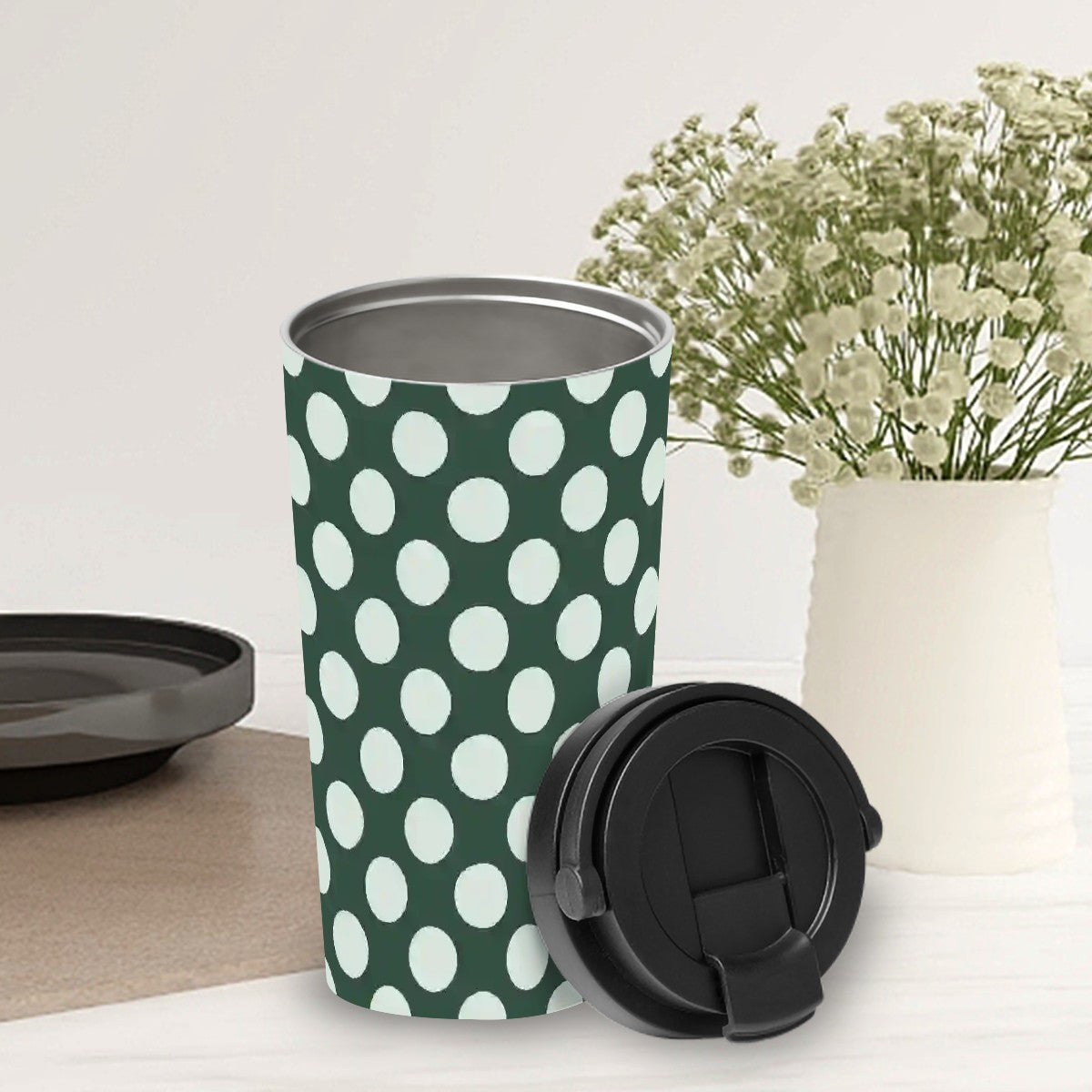 Green travel mug with white polka dots on a light surface with a vase of flowers in the background.
