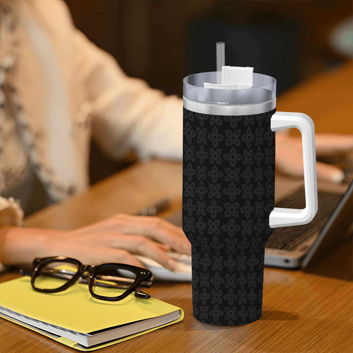 Black patterned tumbler with a white handle on a desk with a person using a laptop in the background.