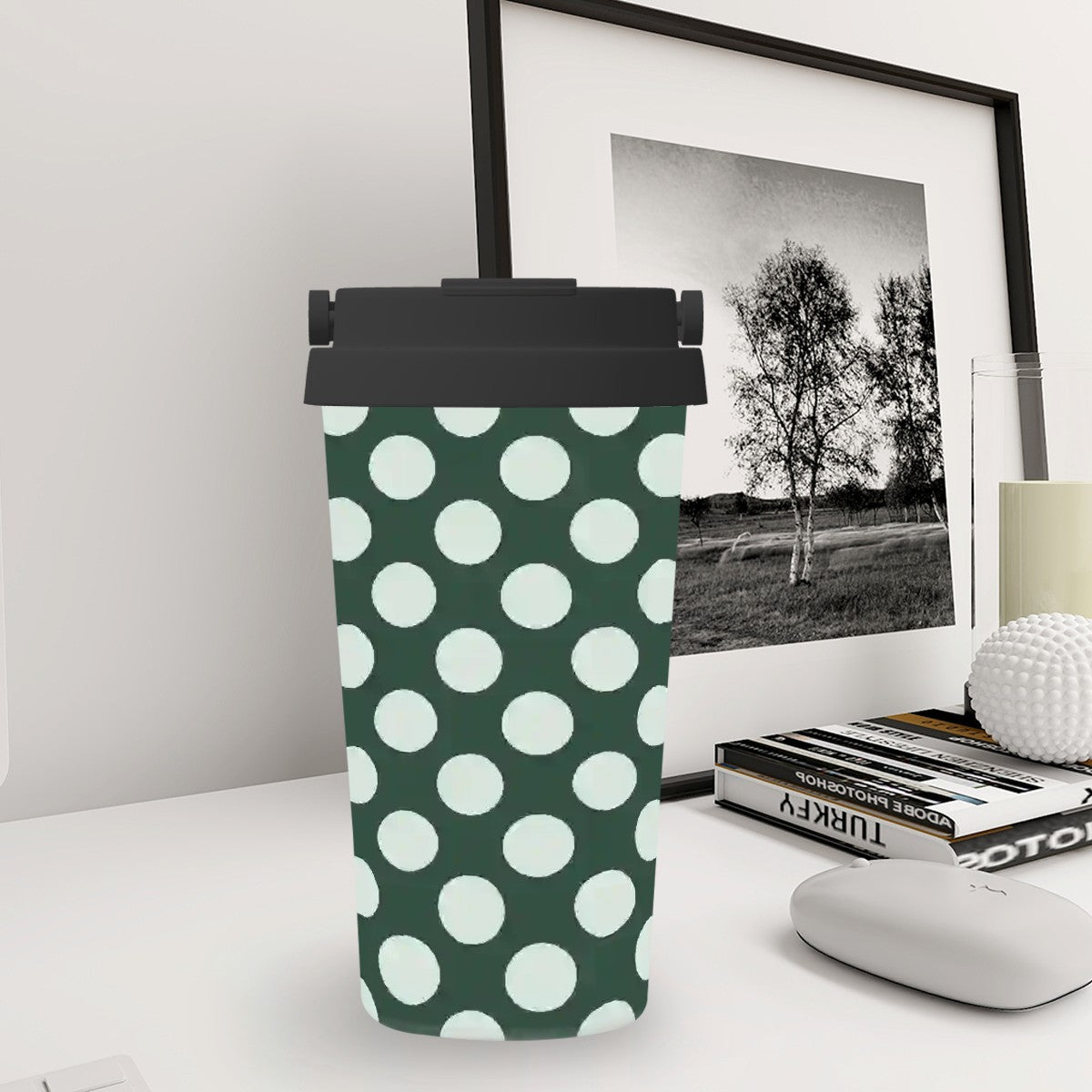 Green tumbler with white polka dots on a desk with books and a framed picture in the background.