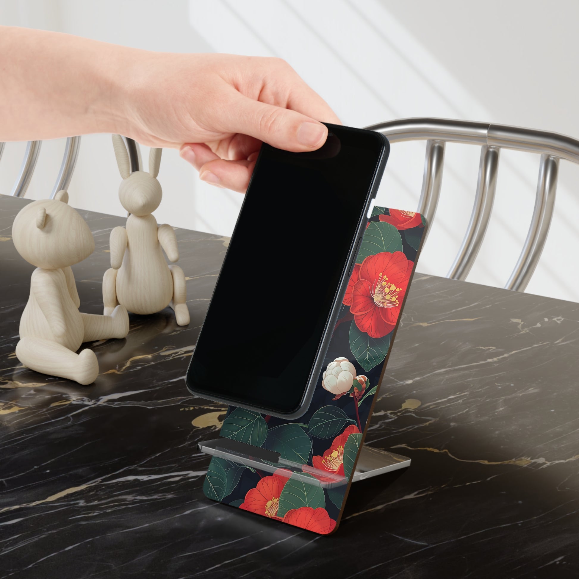 Phone on a floral stand being held by a hand on a marble surface with decorative items.