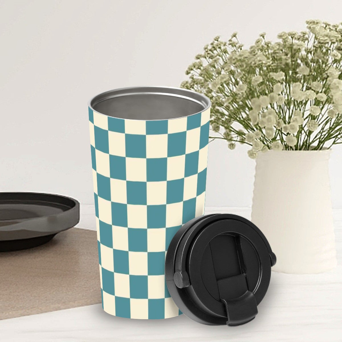 Checkered patterned tumbler with a black lid on a white surface with a vase of flowers in the background.