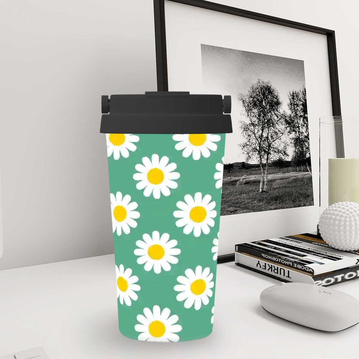 Tumbler with daisy pattern on a desk next to a framed picture and books.