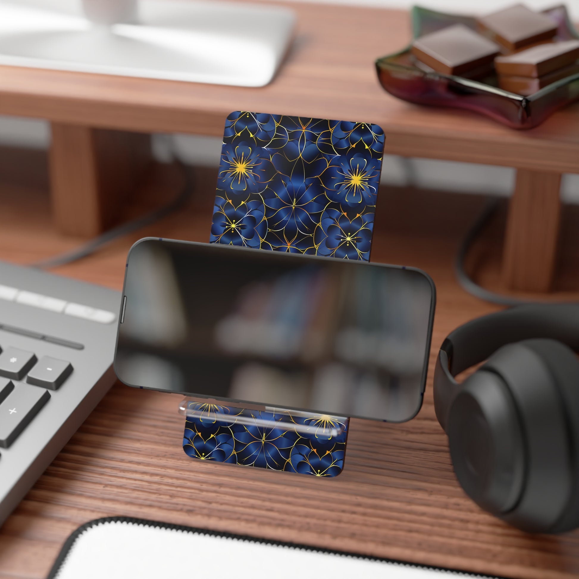 Phone stand with blue floral design on a desk with keyboard, mouse, and headphones.