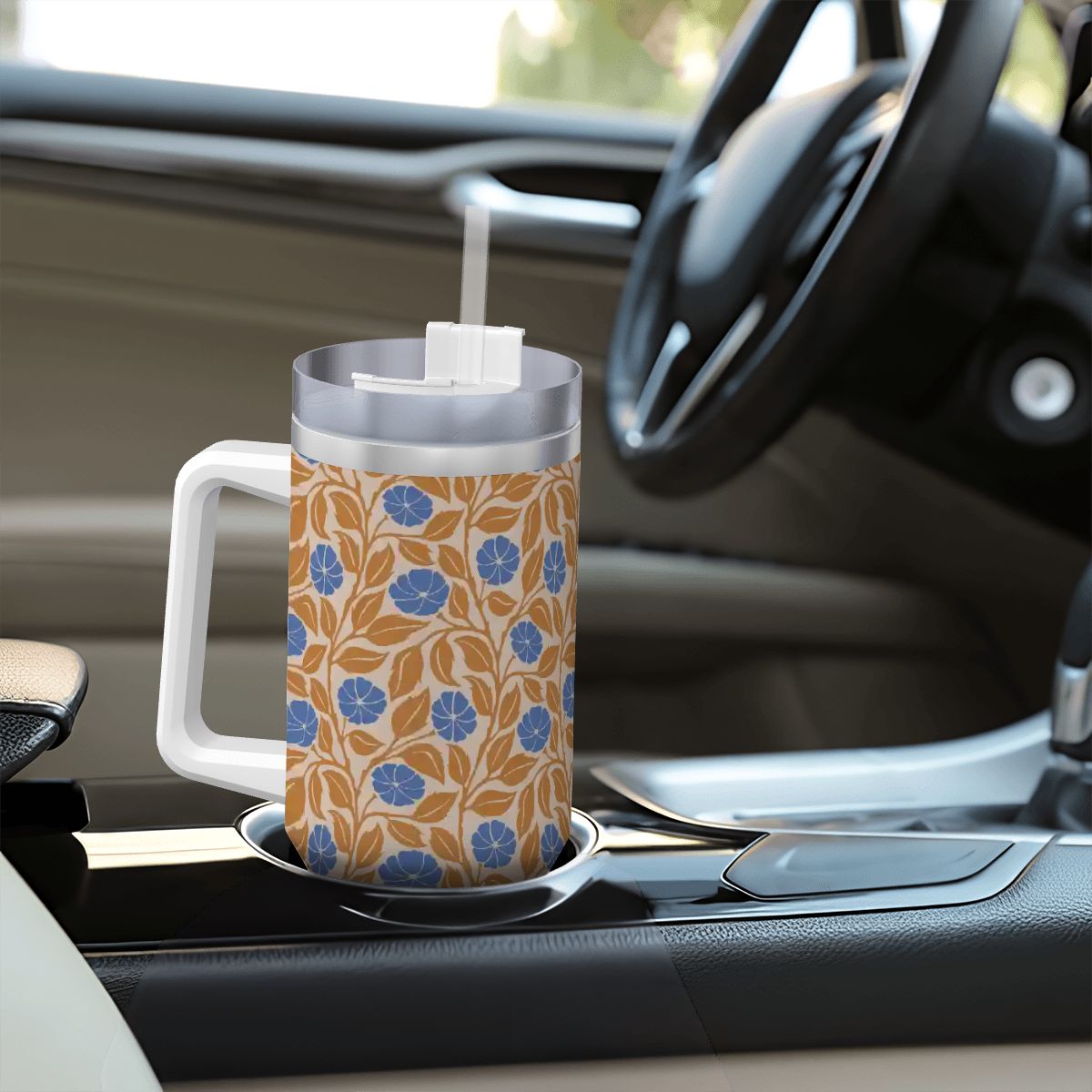 A handled insulated tumbler with a blue floral pattern on a tan background, sitting in a car cup holder. The tumbler has a clear lid with a white straw.