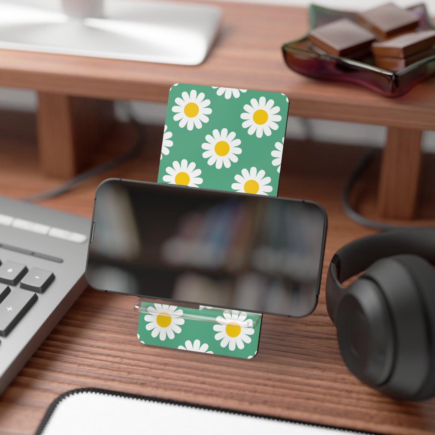 Phone stand with daisy pattern on a desk with keyboard, headphones, and books.