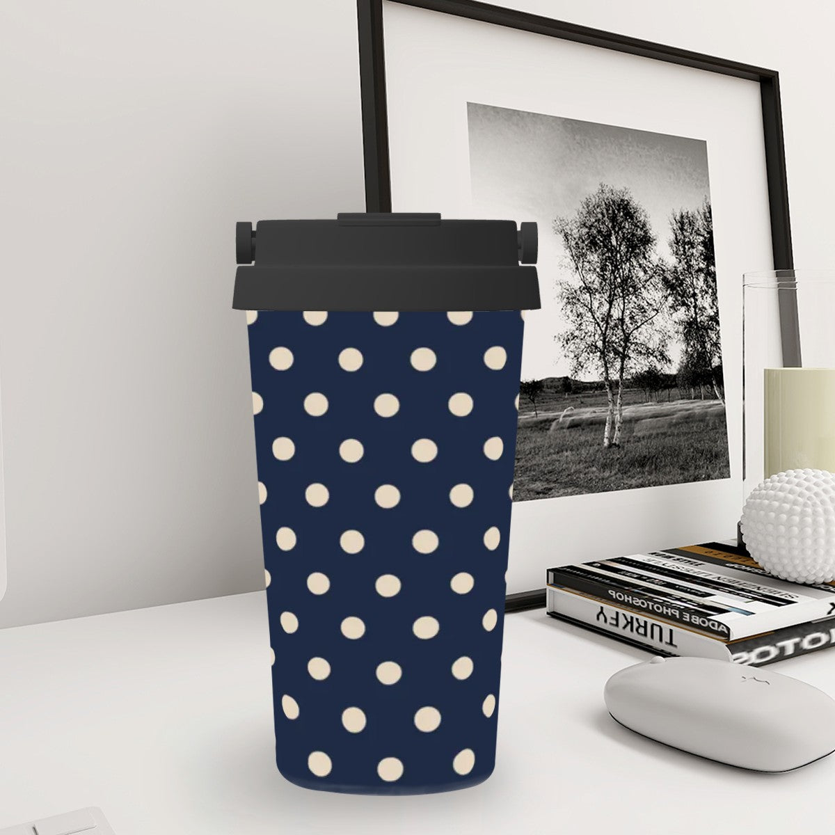 Navy blue tumbler with white polka dots on a desk with books and a framed picture in the background.