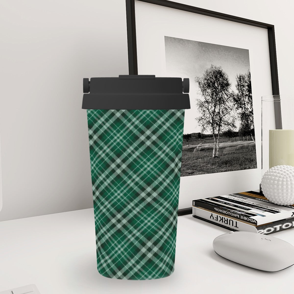 Green plaid tumbler with black lid on a desk next to a framed picture and books.