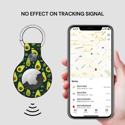A keychain-shaped Apple AirTag blocker with a green avocado print and a silver metal key ring, shown next to a hand holding a smartphone displaying a tracking map. Text at the top reads 'NO EFFECT ON TRACKING SIGNAL'.