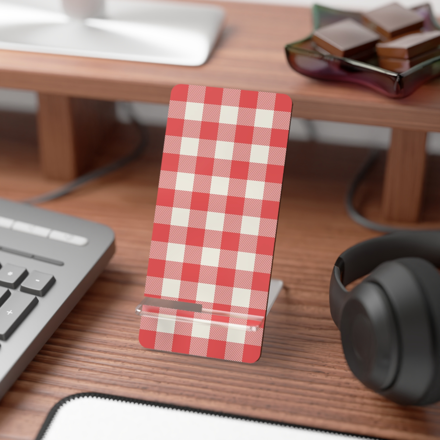 A slim rectangular phone stand or card holder with a red and white gingham check pattern, standing on a wooden desk beside a keyboard and headphones.