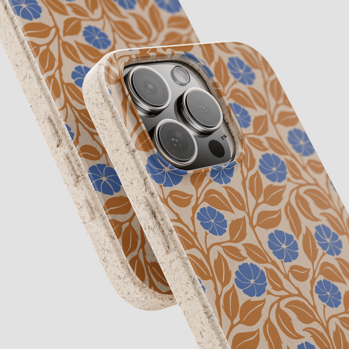 A close-up of a phone case with a beige background, brown leaf pattern, and blue flower motifs, shown overlapping another case on a light gray background.