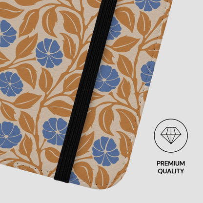 Close-up of a fabric zippered pouch or notebook cover with a cream background, tan leaf pattern, and blue flower motifs, plus a black elastic band. A small 'PREMIUM QUALITY' diamond icon appears on the right.