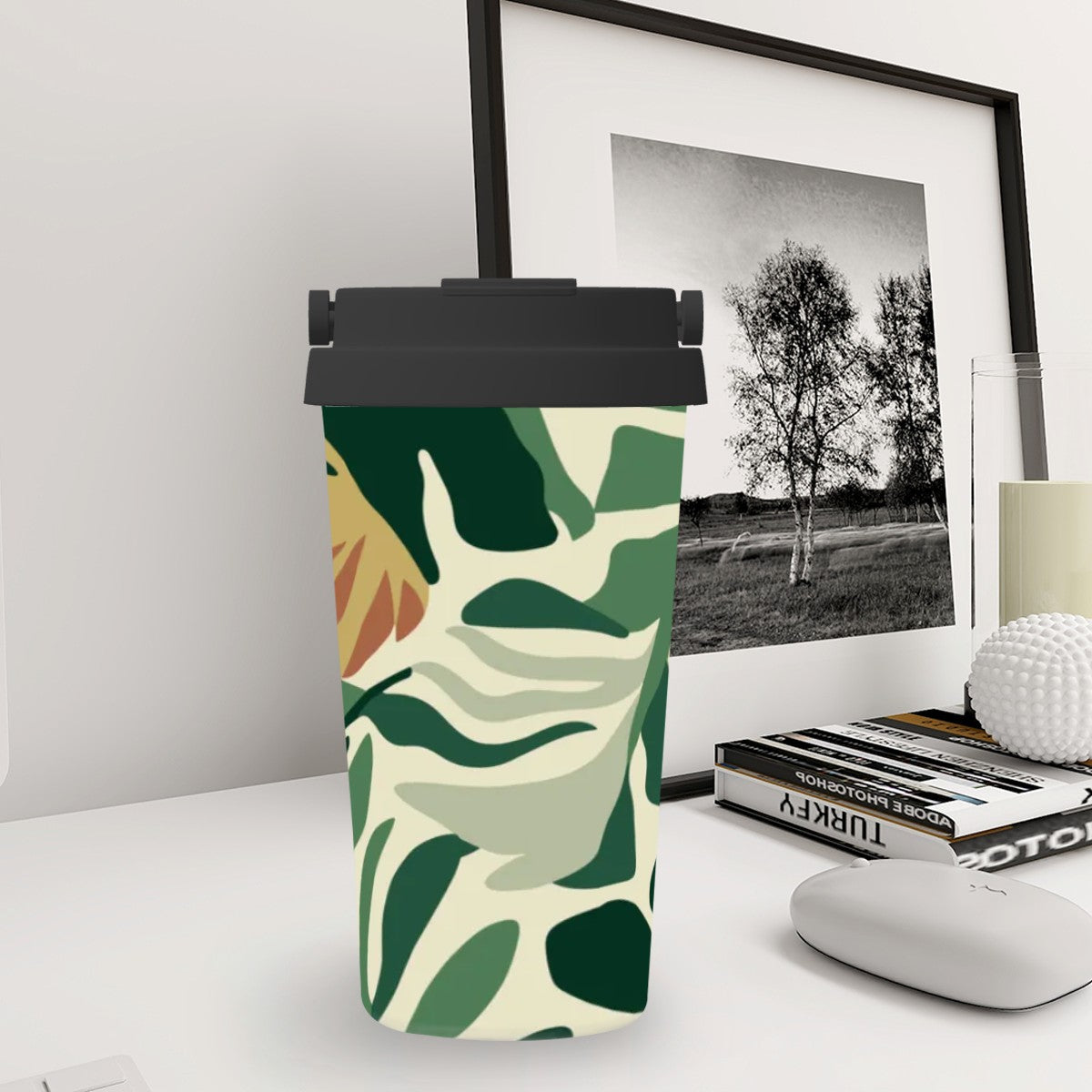 Tumbler with green leaf pattern on a desk with books and a framed picture in the background
