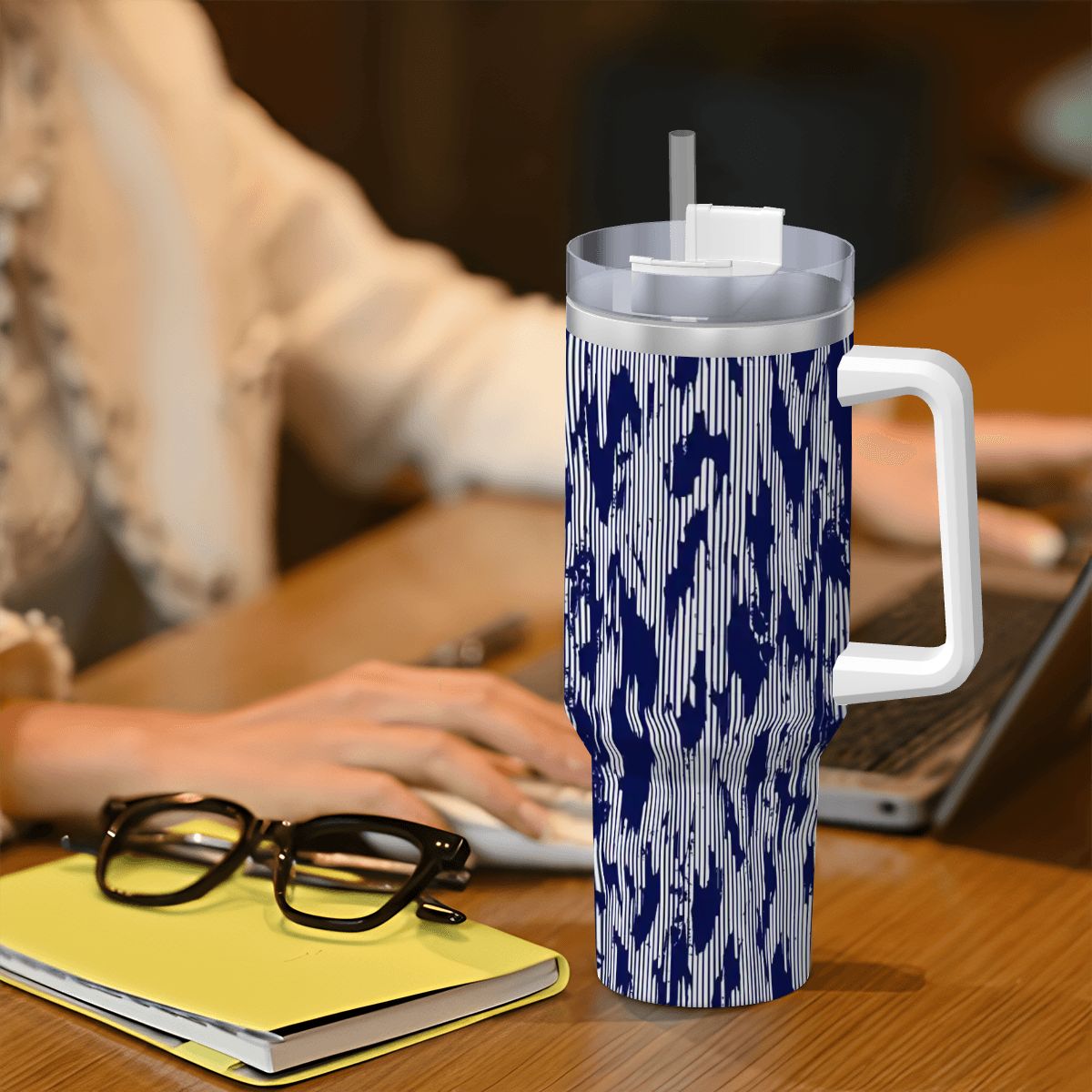 Blue and white patterned tumbler with a straw on a desk next to a person using a laptop.