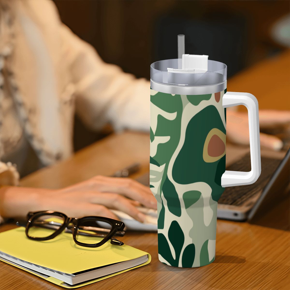 Camouflage-patterned mug with a straw on a desk next to a laptop and glasses