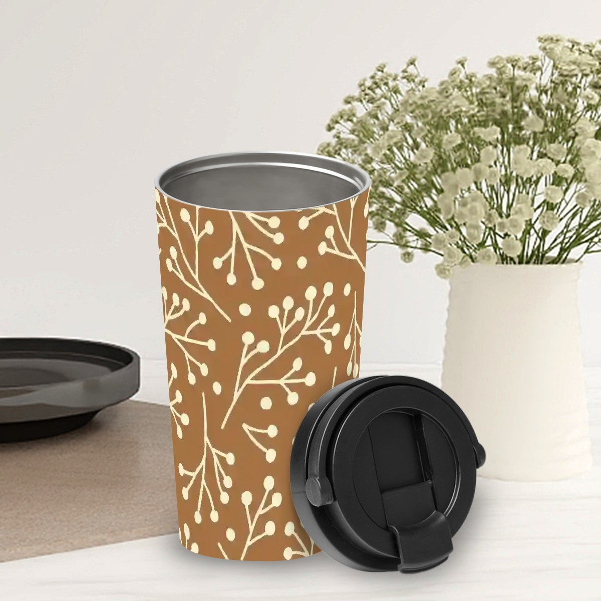 Brown travel mug with white floral pattern on a white surface with a vase of flowers in the background.