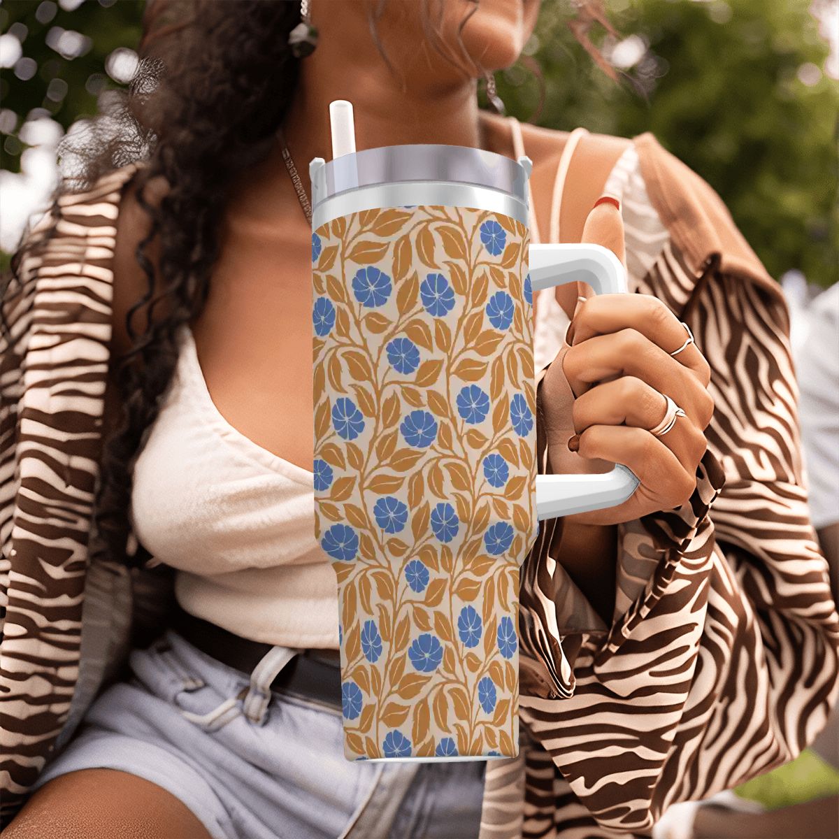 A person outdoors holding a handled tumbler with a beige floral pattern, blue flowers, and a white handle. The person wears a white top, denim shorts, and a brown-and-white zebra-print cardigan.